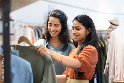 Obraz na plátně Two stylish multiethnic women selecting clothes together in modern boutique, reviewing quality of green garment on hanger, engaged in shopping process, exploring new collection in retailspace