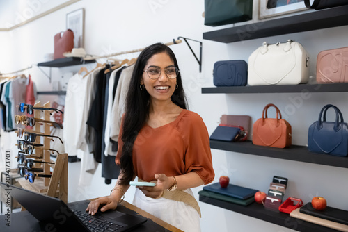Billede på lærred Indian female shop assistant stands behind checkout counter in trendy boutique, holds smartphone in using laptop engaged in positive interaction or completing online order
