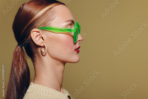Quadro su tela Profile of a young woman with sleek hair tied back wearing green sunglasses and hoop earrings on beige background