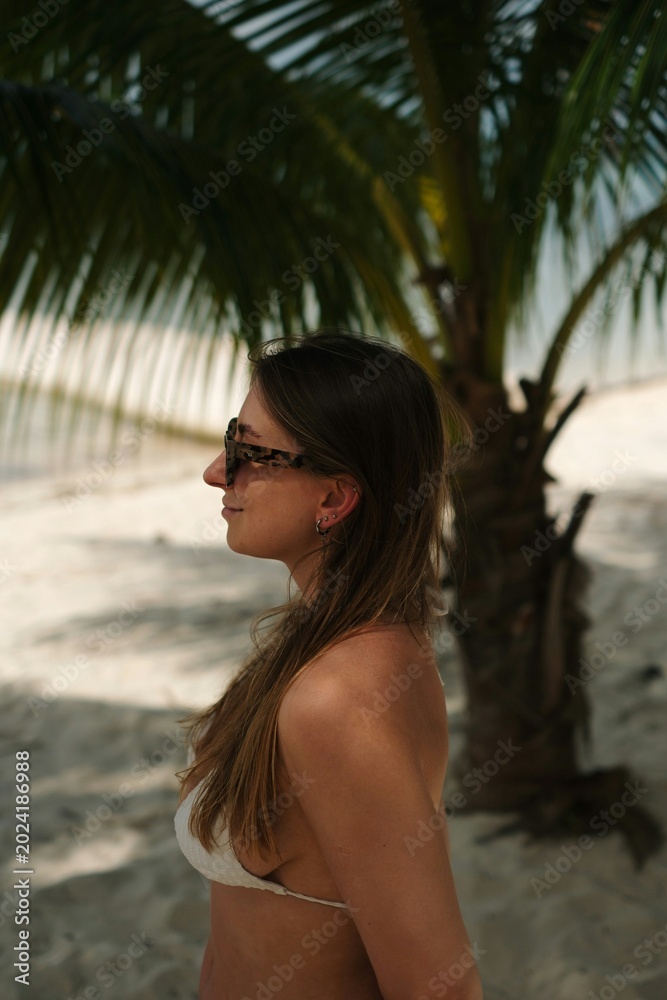 Obraz premium Woman in Bikini on Tropical Beach: A young woman with long brown hair and sunglasses, wearing a white bikini, stands on a sandy beach with a palm tree behind her.
