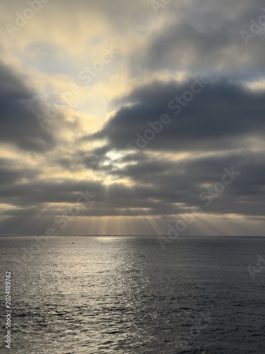 Photography A vertical portrait format composition highlighting sharp, high-contrast dramatic crepuscular light rays streaming down to the coastal ocean waters in La Jolla, California