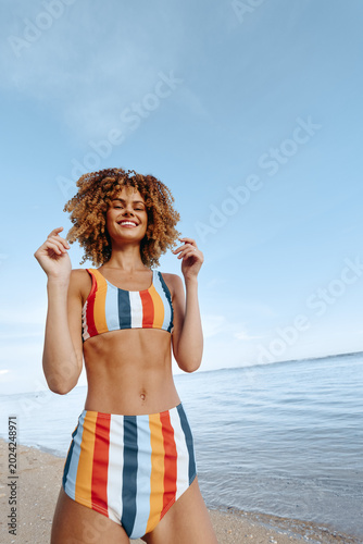 A smiling young woman in a colorful striped bikini stands on a sunny beach  posing confidently. Clear blue sky  calm sea  light breeze  carefree mood  summer vibe  woman enjoying vacation outdoors.