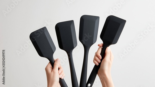 Four black silicone spatulas held by hands against a light gray background, showcasing different shapes and sizes for versatile cooking and baking applications