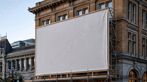 Retail billboard mockup on the facade of a downtown brick building, large format printed graphic on a canvas banner stretching three stories, scaffolding beside it, urban outdoor advertising placeme