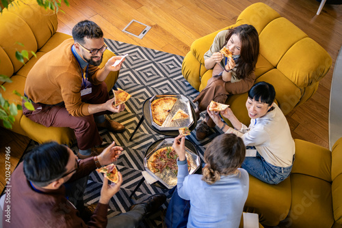 Colleagues having a pizza party, eating lunch together in a modern office lounge area, enjoying a teamwork break