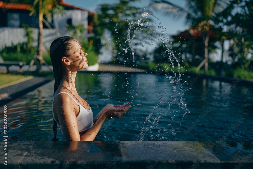 Fototapeta premium Young woman in white swimsuit playing with water in outdoor pool during sunny day. Relaxed lifestyle concept with natural light and tropical background creating calm mood.