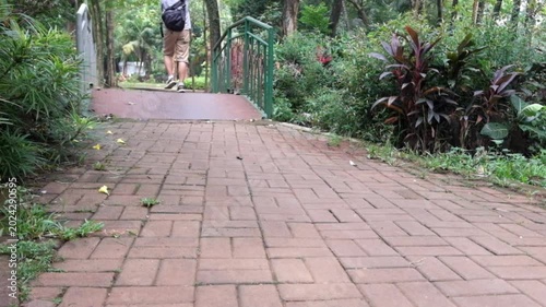 Young man wearing a backpack walks along a paved path in a lush green park setting while moving toward a small metal bridge in this smooth cinematic 4k video clip