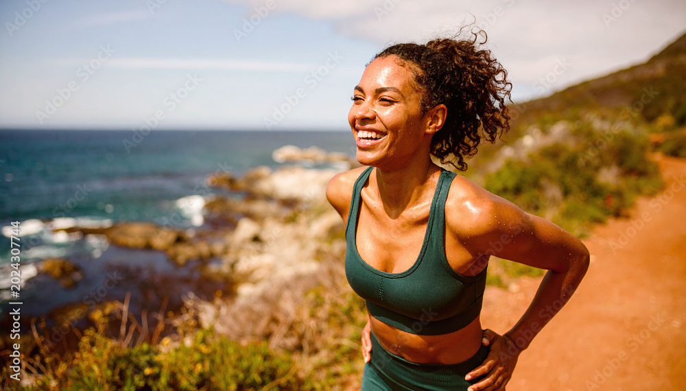 Fototapeta premium Radiant woman in green sportswear smiles after a workout on a scenic coastal path. Capturing fitness, joy, and wellness, this vibrant image is ideal for health and lifestyle campaigns.