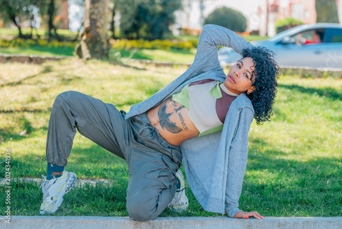 Confident young woman in street-style outfit — cargo pants, cropped hoodie, chunky sneakers — striking a dynamic floor pose in a sunny urban park, showcasing tattoo