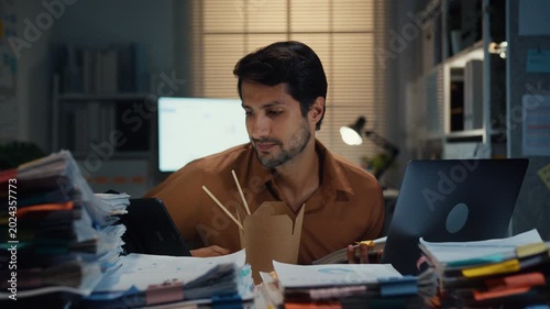 Young Latin businessman taking urgent phone call while reading accounting document on laptop at desk in dark night office. Multitasking overtime, overworked deadline, tired paperwork rush concept.