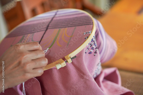 Close-up of artisan hands doing traditional hand embroidery on pink fabric using a wooden hoop, needle, and thread. Concept of craftsmanship, hobby, and handmade textile art.