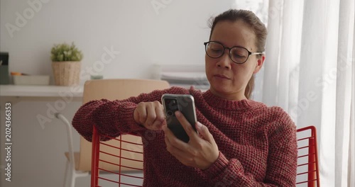 Asian middle woman wear glasses using smartphone on chair by window