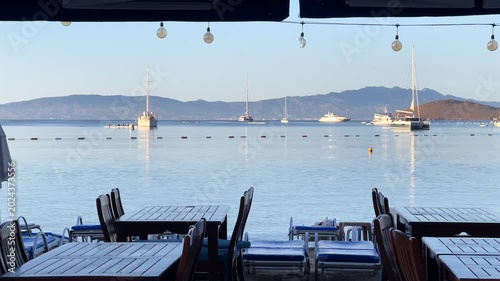 An empty beachfront restaurant terrace in Bitez, Bodrum, looking out over a calm bay filled with anchored yachts. Hanging light bulbs and wooden tables frame a tranquil morning seascape.