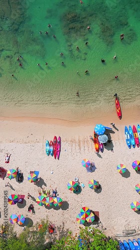 Top down drone view of tropical beach in Phuket, Thailand with colorful umbrellas, kayaks, tourists swimming in turquoise water, and white sandy coastline.