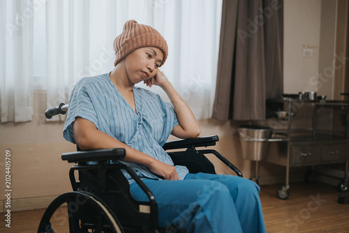 фотографія A melancholic young Asian female patient wearing a pink knit beanie and a blue hospital gown, sitting thoughtfully in a wheelchair inside a hospital room