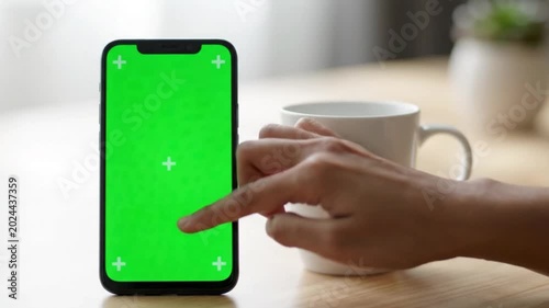 Hand touching a smartphone with a green screen on a wooden table next to a mug