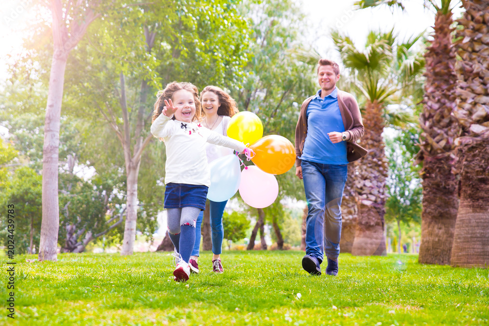 Fototapeta premium A woman and two young girls are running together in a grassy park, smiling and holding colorful balloons on a sunny day. 