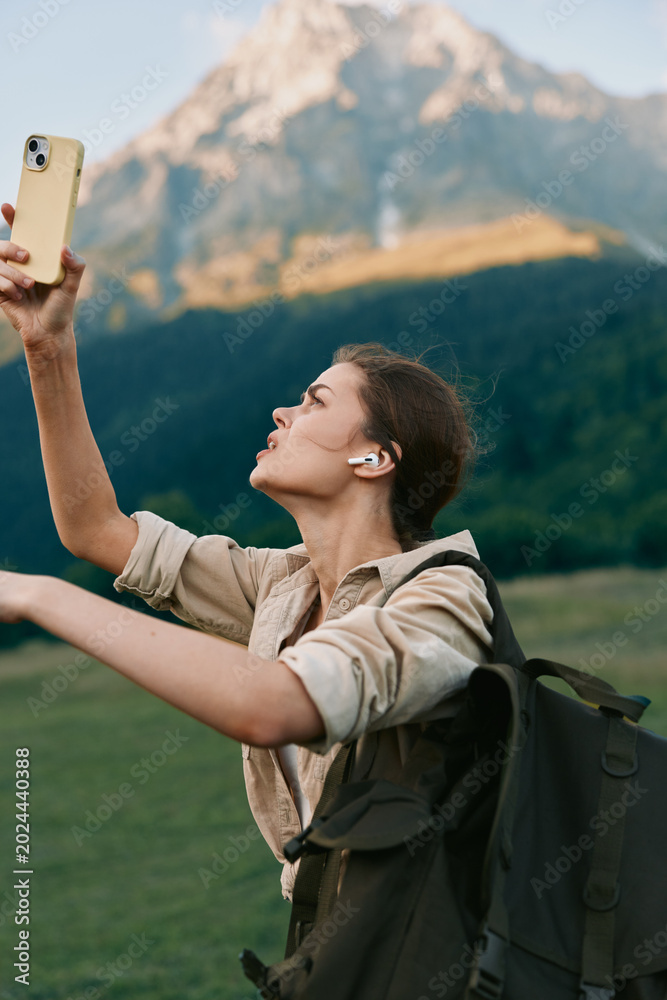 Fototapeta premium Smiling woman with backpack taking a selfie outdoors in front of a dramatic mountain landscape. Casual hiking scene, travel lifestyle, and outdoor exploration with soft natural light.