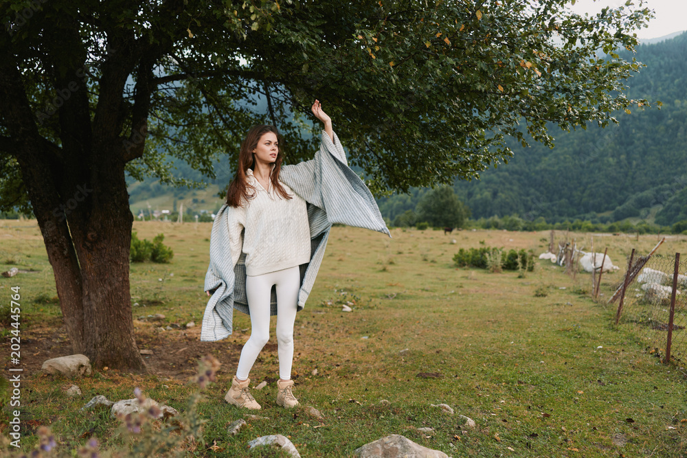 Fototapeta premium A young woman dances in a breezy meadow, wearing white pants and a beige knit sweater with a gray shawl flowing, mountains in the background