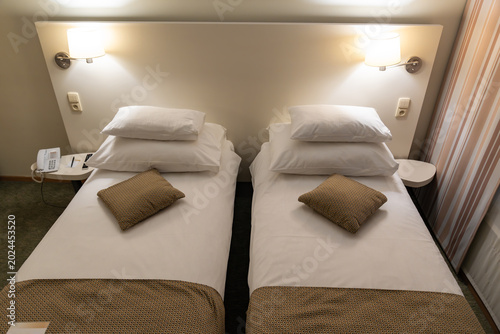 Twin beds with white linens and warm lighting fill a compact hotel bedroom. Neutral furniture and clean arrangement support comfortable overnight accommodation.