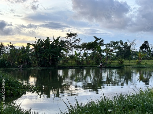 Silhouette of a local fisherman on a small wooden boat in a tranquil tropical river surrounded by lush green vegetation and banana trees under a cloudy sky.