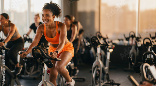 Diverse group of energetic young people laughing and smiling while participating in an intense indoor cycling spin class at a modern fitness gym studio during a golden hour workout session