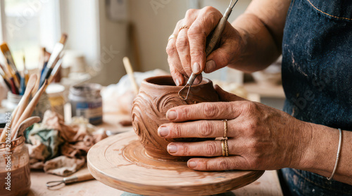 Closeup of artisans hands decorating clay pot with carving tool.