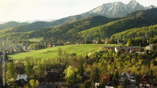 Mountain village houses in green alpine valley during sunrise. Scenic morning landscape with snowy Tatra peak and pine forest in Poland near Zakopane resort area at spring time.