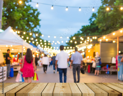 A blurred view of a lively outdoor market scene at dusk, illuminated by strings of warm lights, with a wooden surface in the foreground.