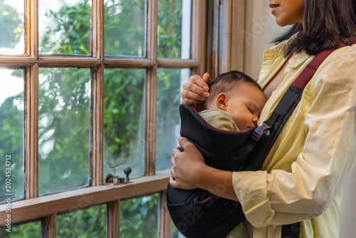 Canvas Print Mother cradling sleeping baby in carrier by window, sunlight activating fit grap