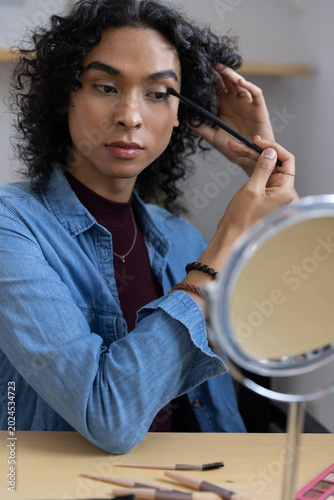 Non-binary adult in denim shirt applying mascara wand while fixing hair at vanity with round mirror