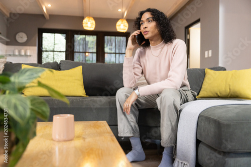 Non-binary person sitting on right of gray sofa at home, talking on smartphone, in pink sweater