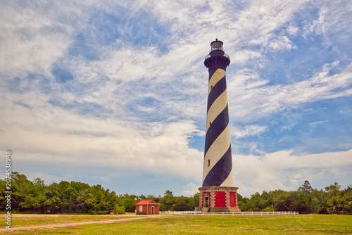 A daytime landscape of the Cape Hatteras Lighthouse on the beach under a cloudy blue sky on the Outer Banks in North Carolina.
