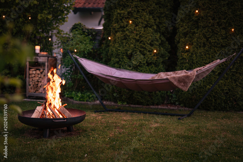 Hammock with fairy lights in garden. Cozy evening atmosphere with a campfire in a fire bowl. Background for leisure activities for outdoors.