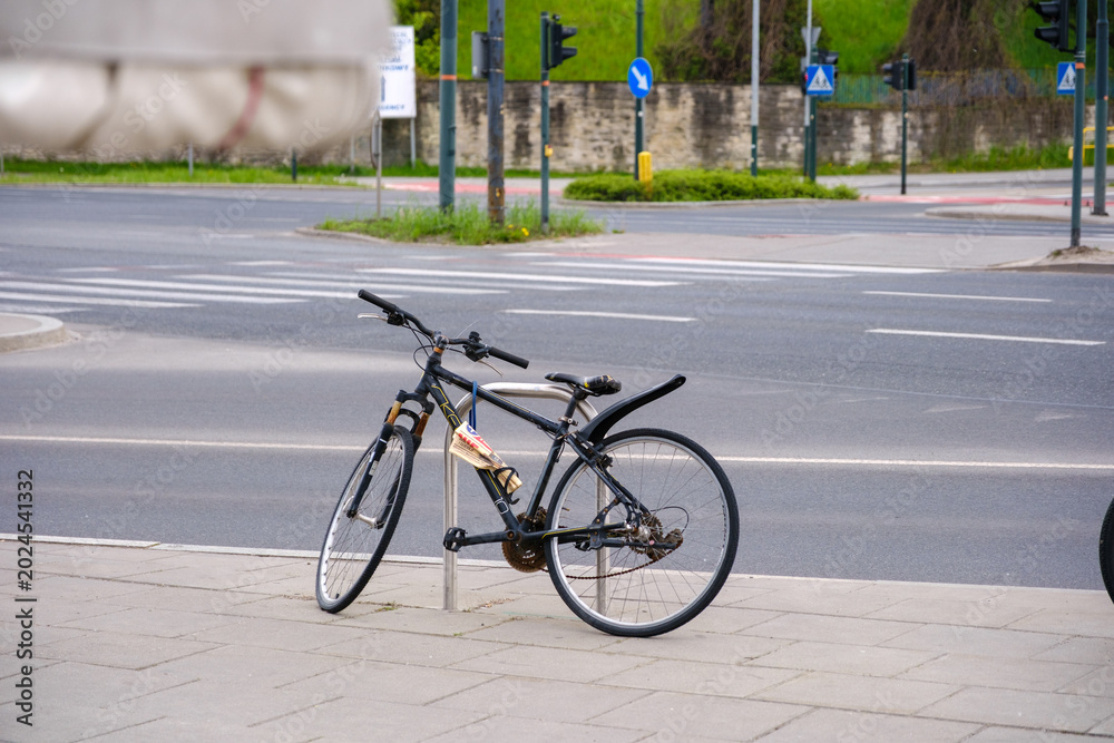 Naklejka premium Black mountain bike secured with a chain lock to a metal post on a city sidewalk, with an empty crosswalk and road infrastructure in the background, symbolizing urban transport and waiting