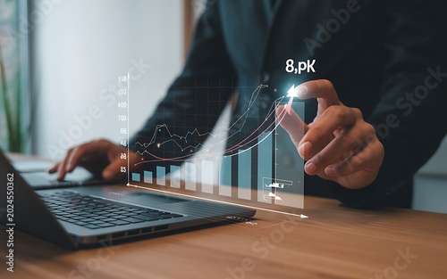 A businessman in a dark suit uses a laptop while a glowing holographic financial graph with bar charts and line graphs rises from the desk, with their finger pointing to an upward trend on the
