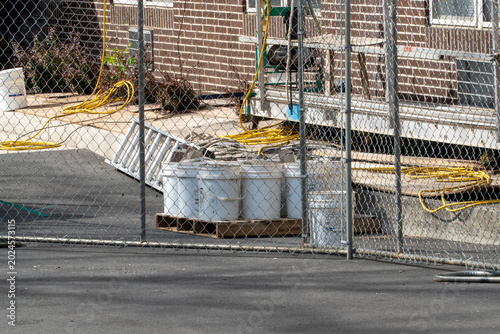 Vászonkép Construction site materials behind a chain-link fence, featuring white buckets, an aluminum ladder, and coiled yellow hoses, depicting a temporary work area or building renovation project