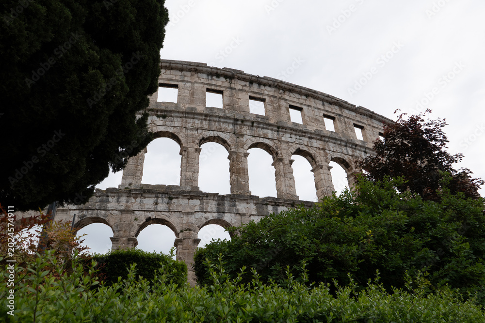 Obraz premium Ancient Roman arena amphitheater in Pula Croatia surrounded by green trees under dramatic cloudy sky view
