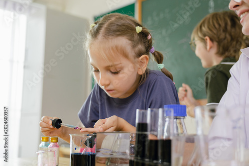 Obraz na plátne A young girl carefully conducts a science experiment with liquids in a classroom, focusing intently while a teacher and another student observe in the background