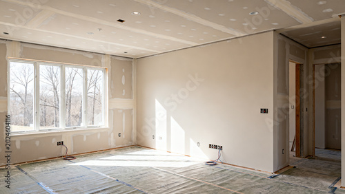Interior of a new house under construction, showing unfinished walls, ceiling, and a large window with natural light streaming in, highlighting the home renovation process