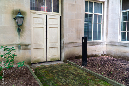 Foto Historic stone building entrance with weathered double doors, vintage verdigris lantern, mossy brick path, and reflective windows showing an 'EXIT' sign, evoking a timeless academic atmosphere
