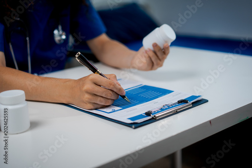 Healthcare professional in scrubs with stethoscope filling out medical forms on clipboard at clinic desk, holding phone; close-up, blue tones, copy space.