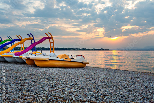 Colorful pedal boats lined up on a pebbled beach at Lago Garda, Italy, with a serene sunset reflecting on the calm water and clouds in the sky