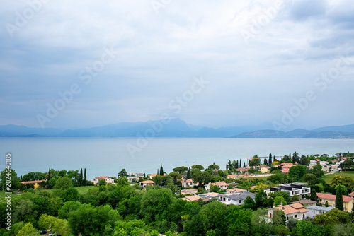 Картината върху платно Scenic landscape of Lago Garda in Italy with tranquil waters, rocky shore, trees, and a charming town illuminated by From above, in the rays of the sun against a blue, cloudless sky