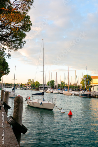 Sailboats moored in a tranquil harbor at Lago Garda, Italy, with lush greenery and a clear sky reflecting on the water's surface