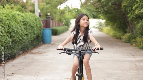 A happy young Asian girl smiles while riding a bicycle on an outdoor path. Representing a healthy lifestyle, active childhood, freedom, and joyful outdoor recreation on a sunny day.