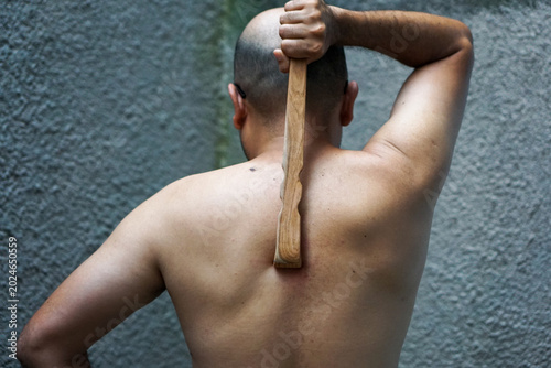 Man holds wooden back scratcher tool behind his back. He prepares for a physical therapy session. The tool aligns with his spine, suggesting self-care. His posture shows focus, not pain