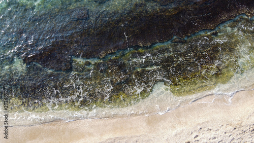 Top-down view of gentle ocean waves washing over a sandy beach and dark rocky reef, showing clear turquoise water and white sea foam in a serene coastal scene.