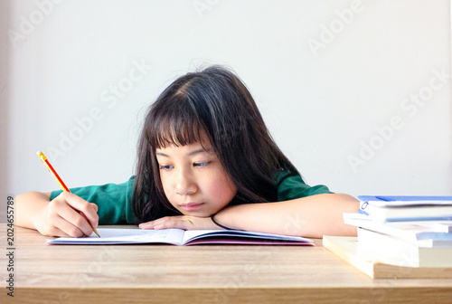 Focused young Asian girl studying at home, resting her arm on a wooden desk while diligently writing in a notebook with a pencil. Primary school student concentrating on homework.
