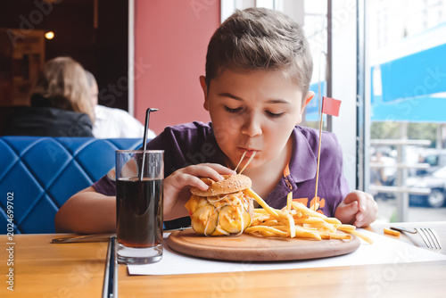 Funny caucasian preteen kid enjoying a juicy hamburger and french fries in a restaurant, childhood nutrition and fast food menu concept.